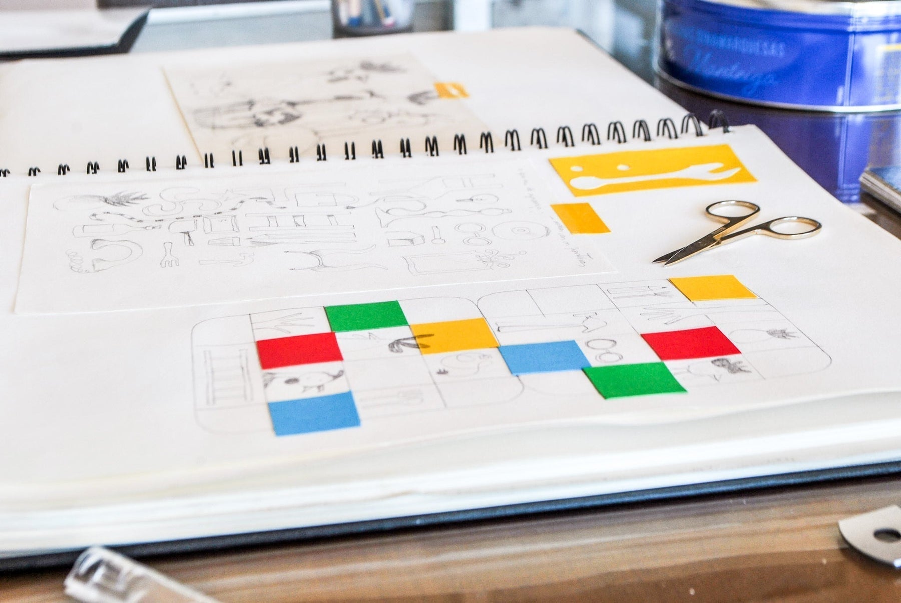 Caderno de páginas brancas aberto sob uma mesa de madeira. As páginas contém rascunhos em grafitti e quadrados de papel amarelo, vermelho, azul e verde compõem uma colagem